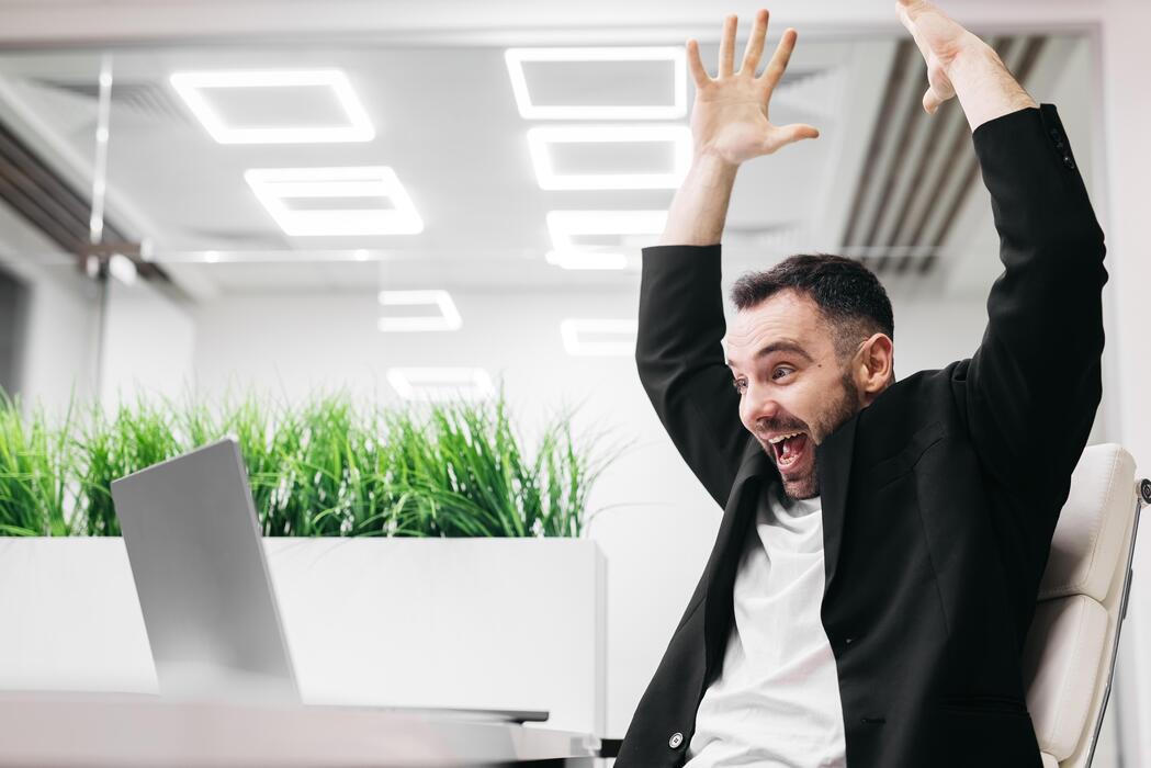 Small business website design free - Male office worker in a black blazer is celebrating with raised arms in a modern workspace with greenery and bright lighting during a successful moment at his desk