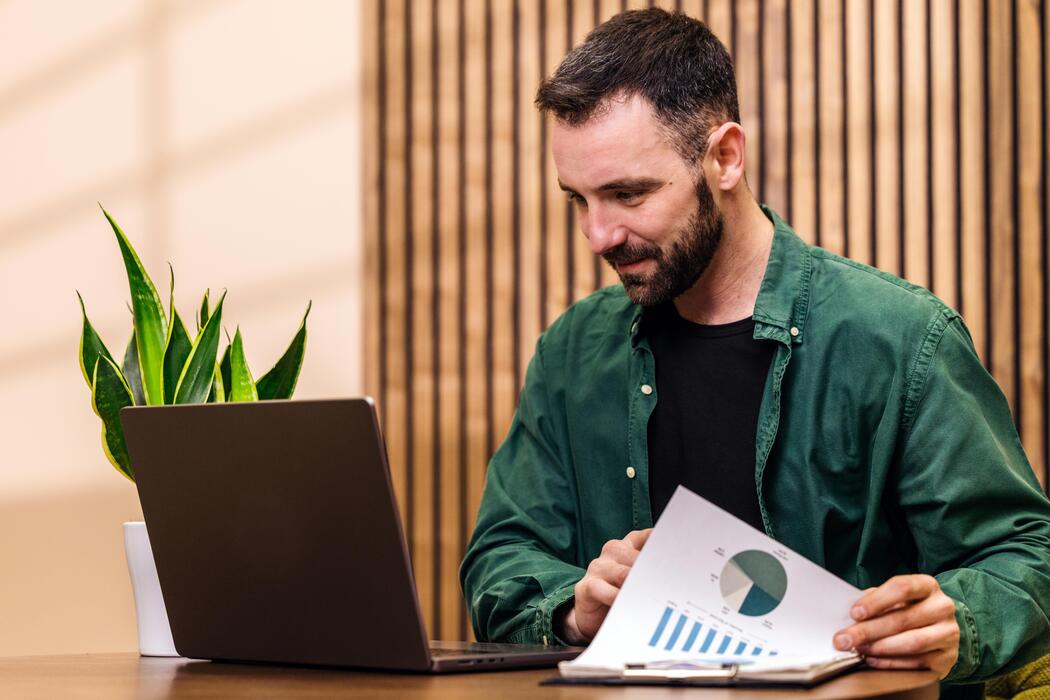 Best website builder for small business free - Male professional in green shirt analyzes financial report with laptop on wooden desk, indoor setting featuring potted plant and wooden wall in the background