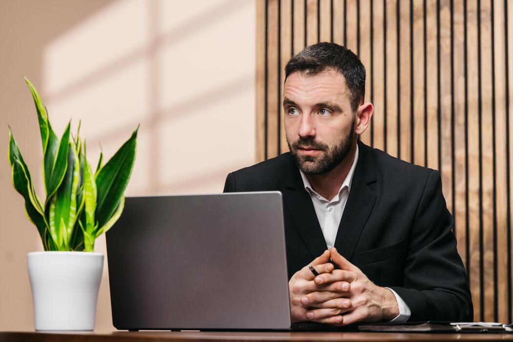 Best website builder for small business free - Businessman with beard in black suit sitting at desk with laptop and potted plant, wooden wall in background, focused expression in modern office setting