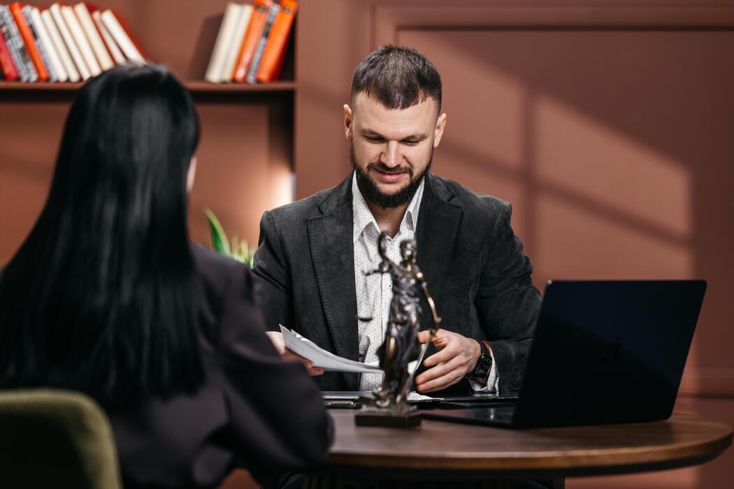 seo basics for small businesses uk - Male lawyer discussing legal documents with a female client in an office, Lady Justice statue placed on the table, bookshelves visible in the background