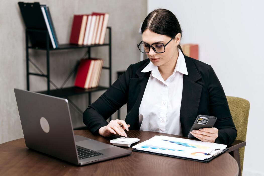 how much does a small business website cost uk - Businesswoman in black suit using smartphone and laptop while reviewing documents on desk with bookshelves in the background