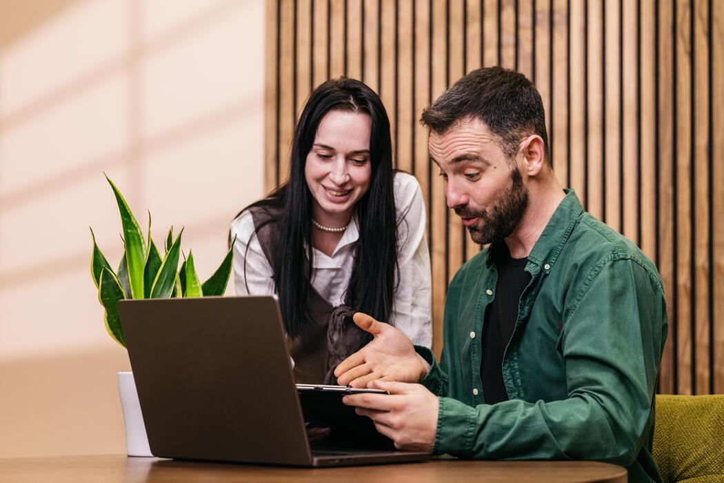 How can I build a website for my small business? - Two professionals, a woman and a man, engaged in discussion while looking at a laptop on a table, with indoor plants and wooden wall in the background