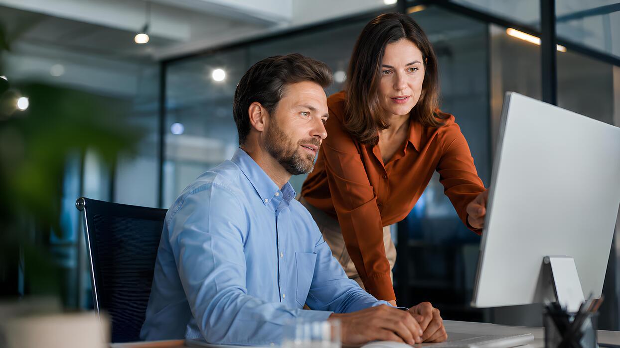 small business website design and hosting - Professional businesspeople collaborate on a computer screen in a modern office workspace. A man sits and a woman stands behind him, smiling as they discuss a company project