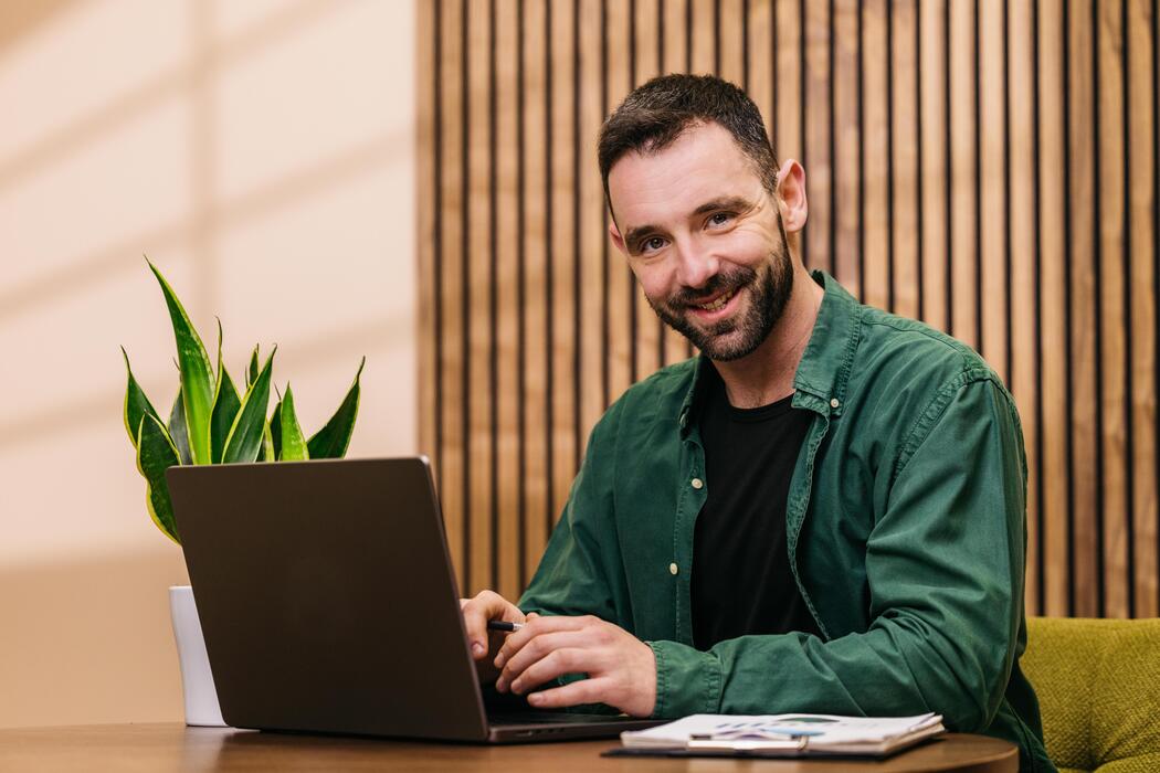 Best small business website design - Male professional with beard wearing green shirt works on laptop at wooden desk with potted plant and notepad in modern office interior