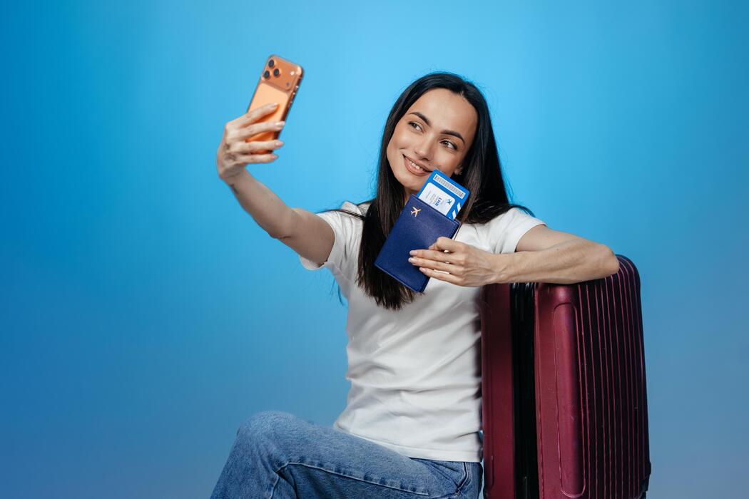 Superkings cigarettes duty free - Young woman taking a selfie with smartphone while holding a passport and boarding pass next to a purple suitcase against a blue background