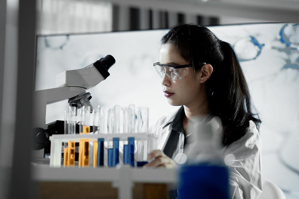 Cheapest research peptides - woman wearing a lab coat and goggles is looking through microscope.
